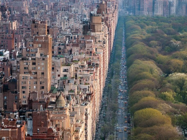 The division of the East and West Aerial view of a city with tall buildings beside a lush green park and tree-lined avenue.